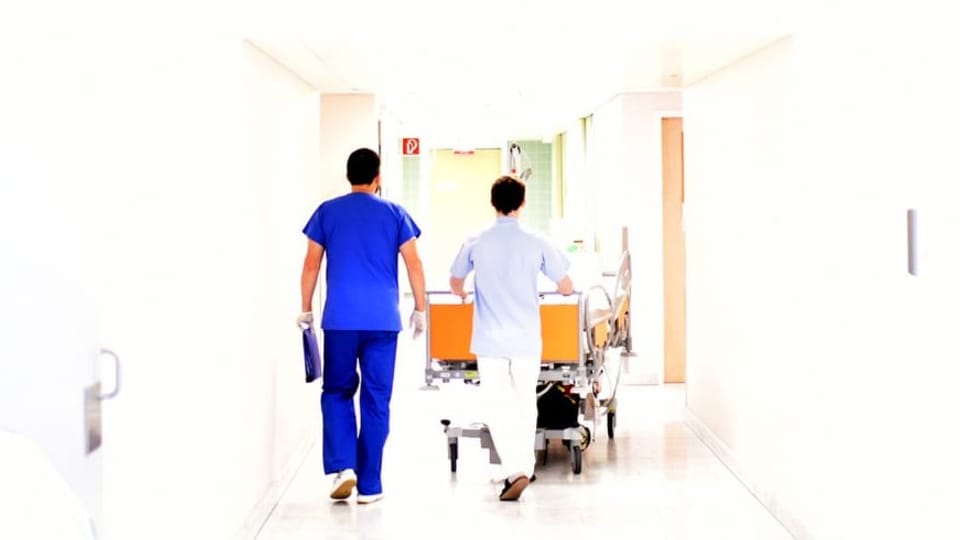 Zwei Spitalmitarbeiter laufen einen Gang hinunter und schieben ein Patientenbett vor sich her.