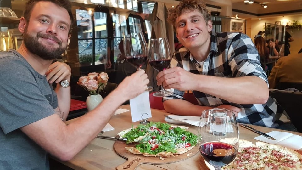 Zwei junge Männer sitzen an einem gedeckten Tisch im Restaurant und stossen mit Rotweingläsern in der Hand an.
