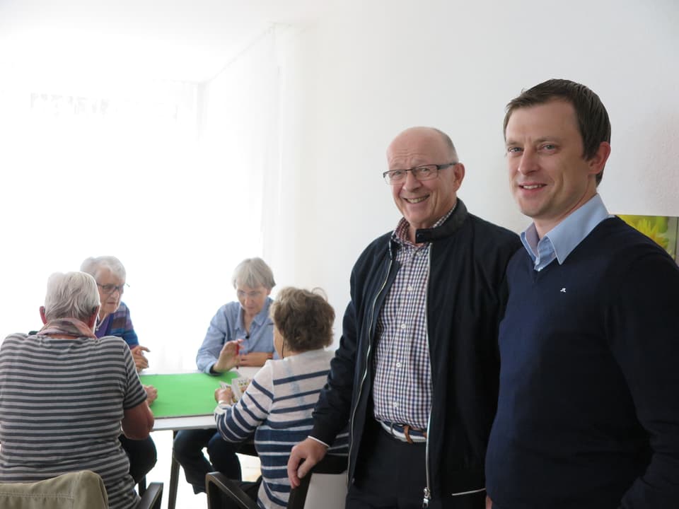 Zwei Vertreter der Wohnbaugenossenschaft vor einer Runde jassender Senioren in einem Gebäude der Siedlung Neuhushof.