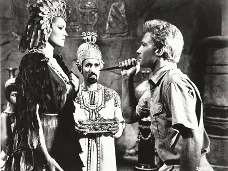 Schwarzweissaufnahme: Ursula Andress als altertümliche Königin wird von John Richardson als jungem Archäologen mit einem Messer bedroht. Im Hintergrund Christopher Lee als Hohepriester mit goldenem Kopfschmuck.