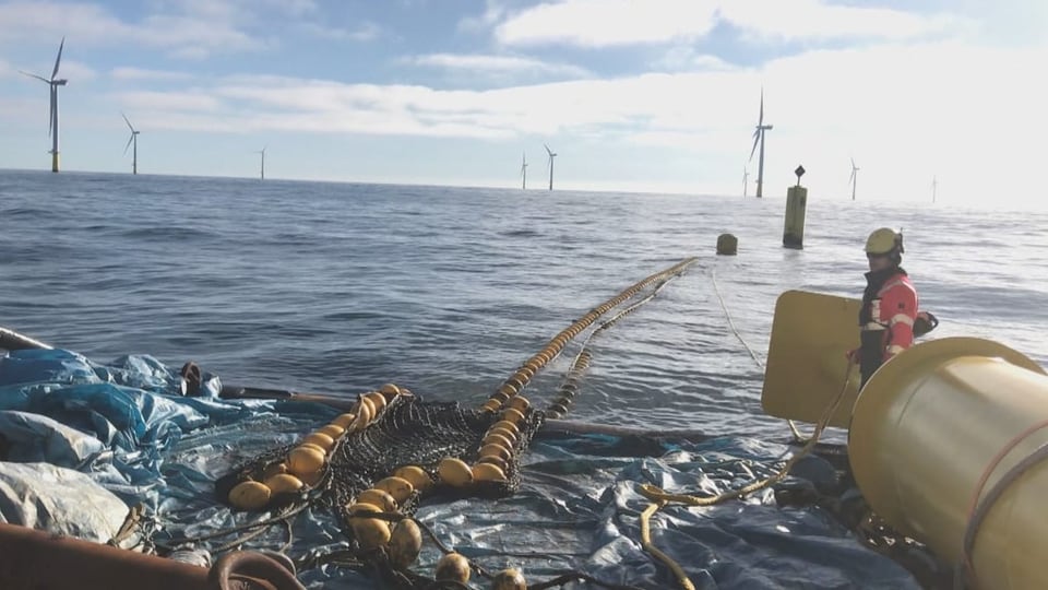 Rechts ein gelbes Rohr und ein Mann in oranger Weste, in der Mitte ein Seil mit gelben Kugeln. Im Hintergrund Windräder.