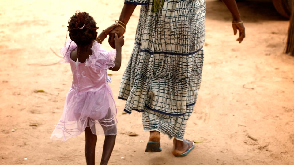 Frau in Afrika führt kleines Mädchen in rosa Kleidchen an der Hand.