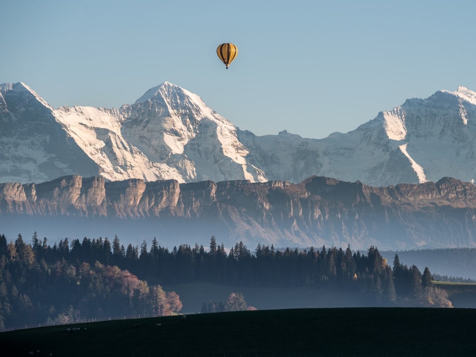 Im Vordergrund ein Hügel im Schatten, dahinter ein bewaldeter Hügel, dahinter Voralpenkrete, dahinter die verschneiten Berner Riesen. Am Himmel steht ein Heissluftballon.