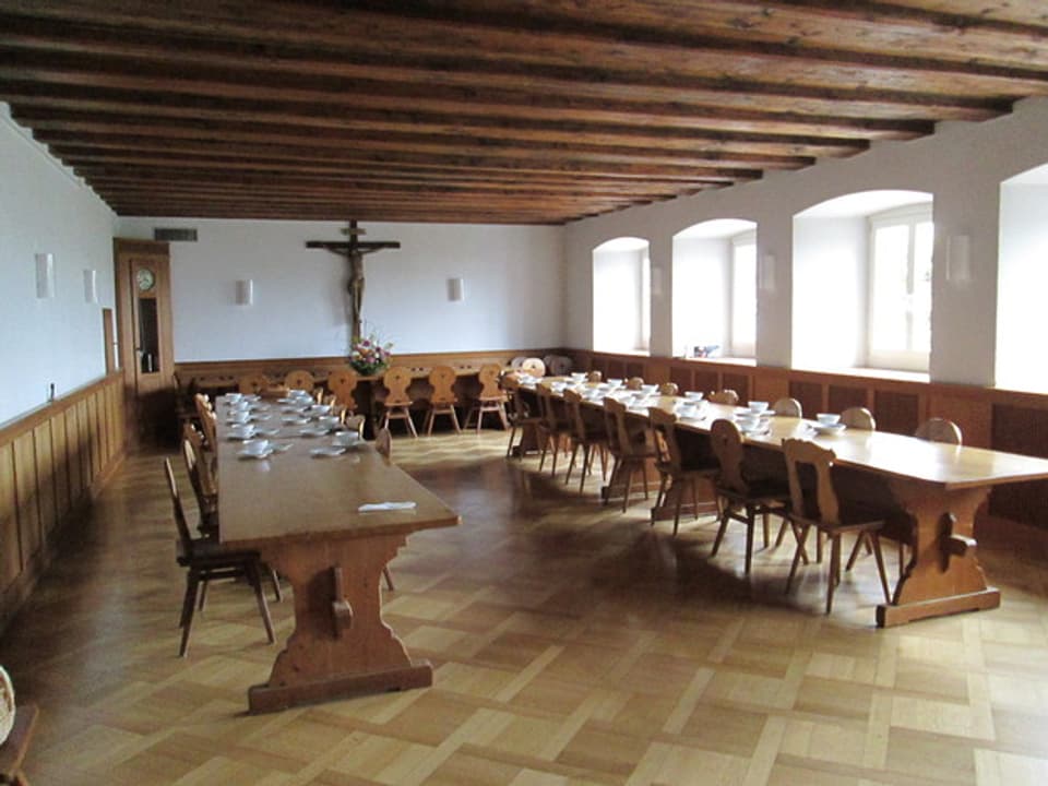 Grosser Raum mit hölzernem Mobiliar, dominiert von einem grossen Kreuz.