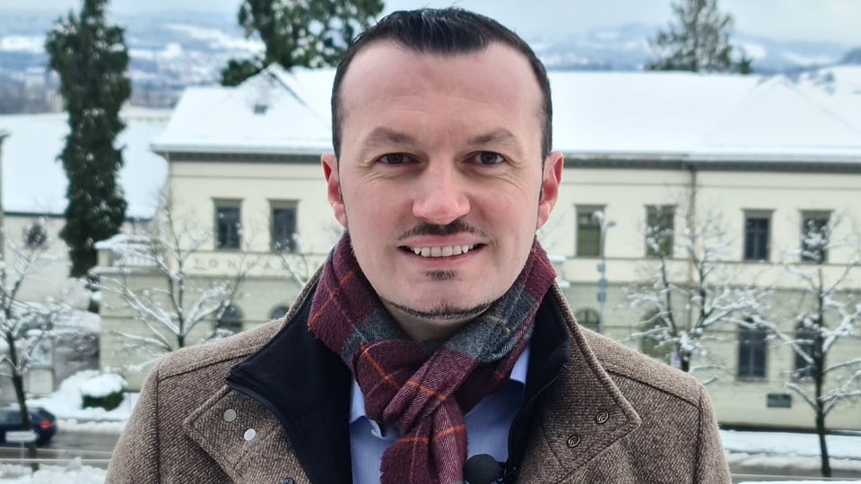 Ein Mann mit einem Schal steht vor einer Schneelandschaft und schaut in die Kamera.