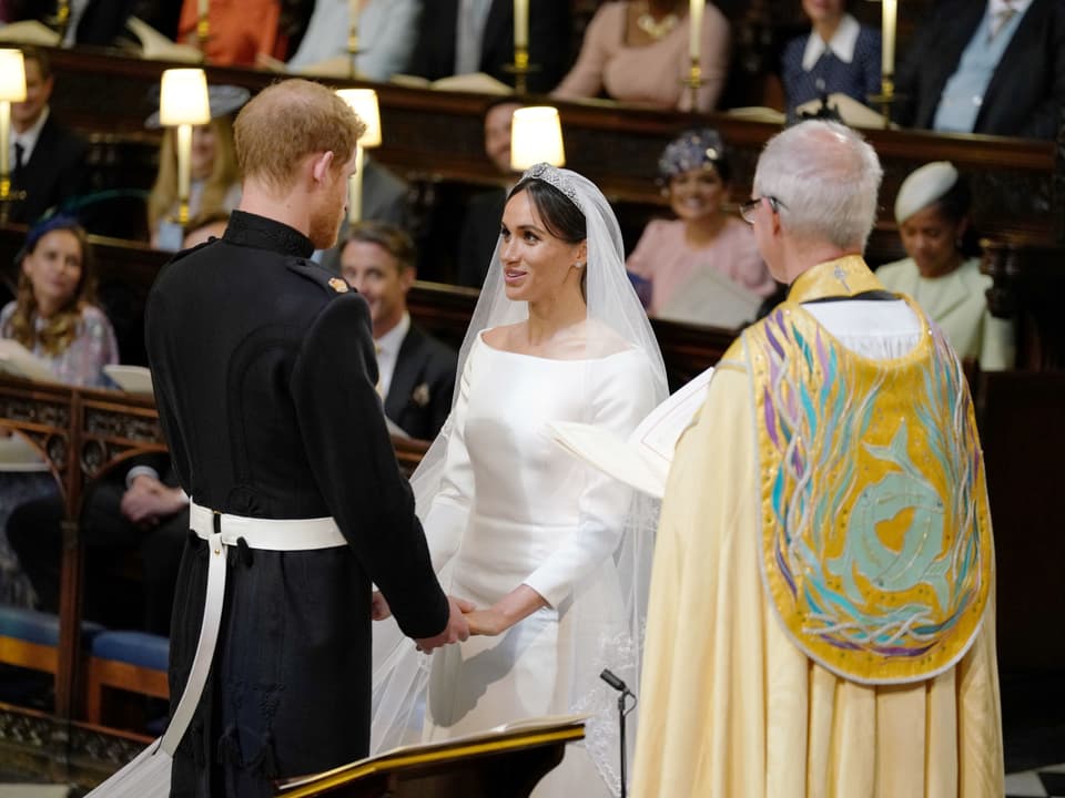 Prinz Harry und Meghan Markle halten beide Hände und stehen vor dem Pfarrer