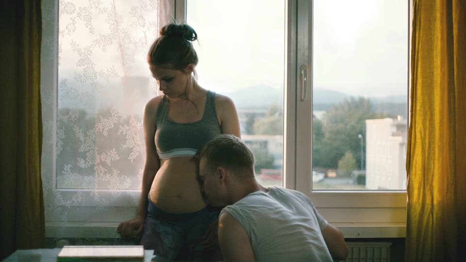Eine schwangere junge Frau mit nacktem Bauch. Ein junger Mann hält sein Gesicht an den Bauch.