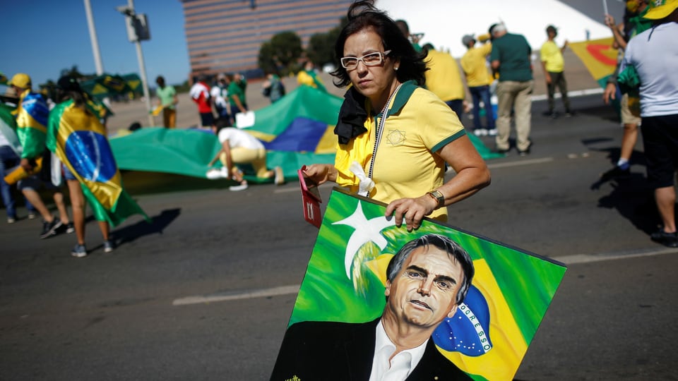 Frau in gelb-grünem Fussballtrikot der  Seleção mit einem gemalten Porträt-Bild Bolsonaros, dahinter weitere Menschen in Fussballshirts.