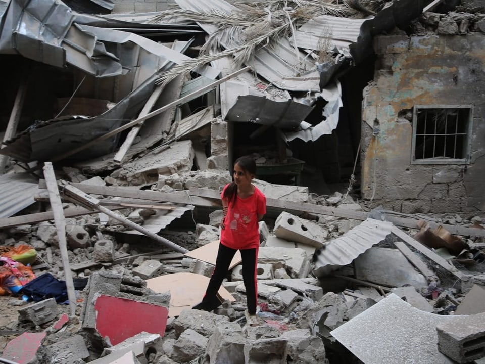 Mädchen in rotem T-Shirt steht inmitten von Trümmern eines zerstörten Gebäudes.