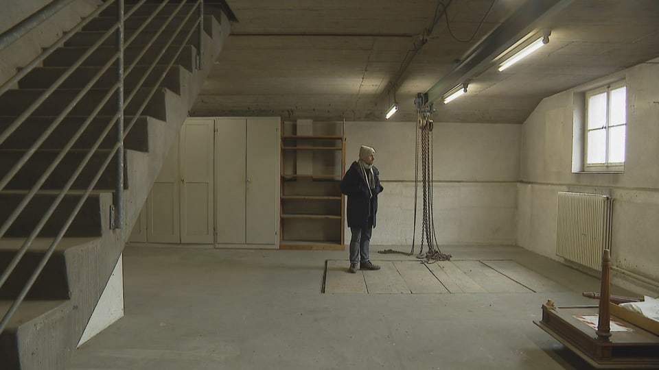 Ein Mann mit Kappe, Schal und Mantel steht in einem leeren, industriell aussehenden Raum. Eine Treppe führt nach oben.