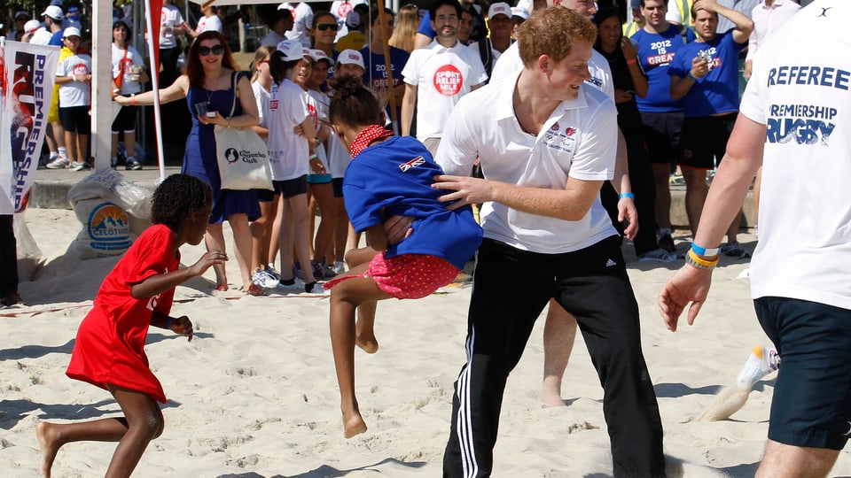 Prinz Harry hebt während dem Rugby-Spiel am Strand ein Mädchen hoch.
