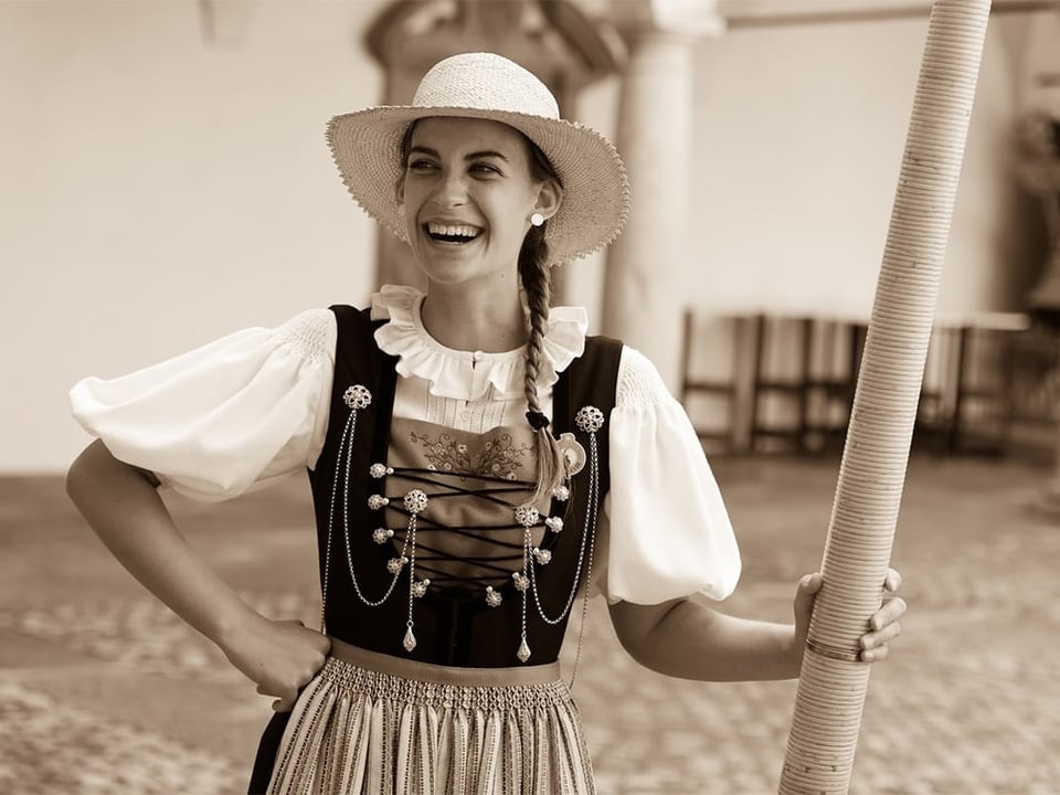 Schwarz-Weiss-Fotografie von einer jungen Trachtenfrau mit Strohhut und Alphorn.