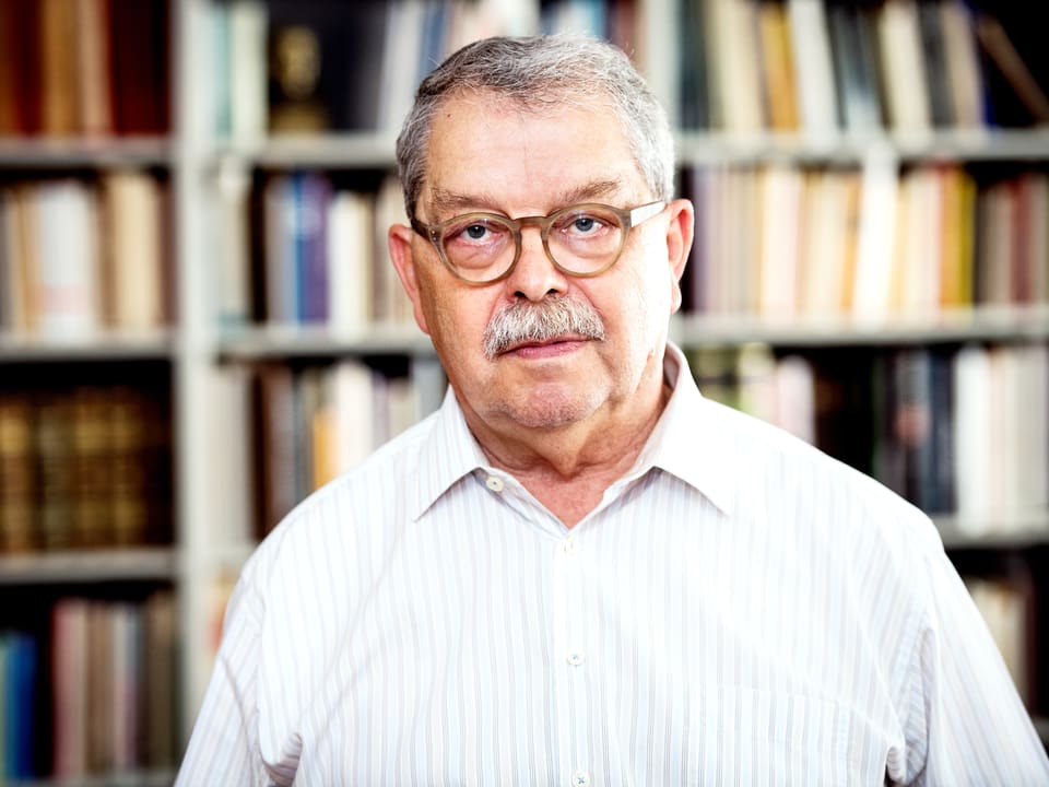 Ein älterer Herr mit Schnauz und Brille steht vor einem Buchregal.