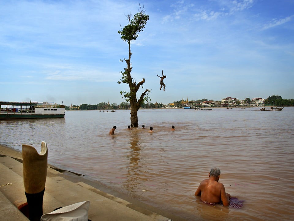 Mann sitzt im Fluss, dahinter springen Kinder von einem Baum ins Wasser
