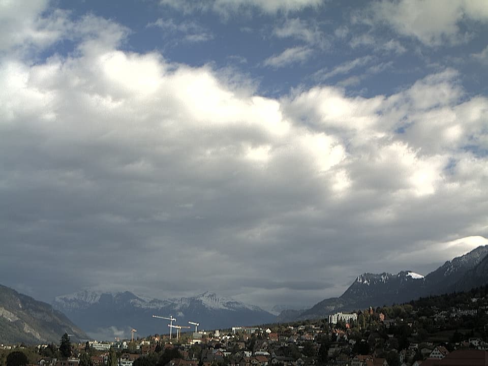 Unten wird die Stadt Chur von der Sonne besonnt. Am Himmel neben vielen grauen Wolken eine grössere Wolkenlücke mit blauem Himmel. Hinten im Tal verschneite Bergen.