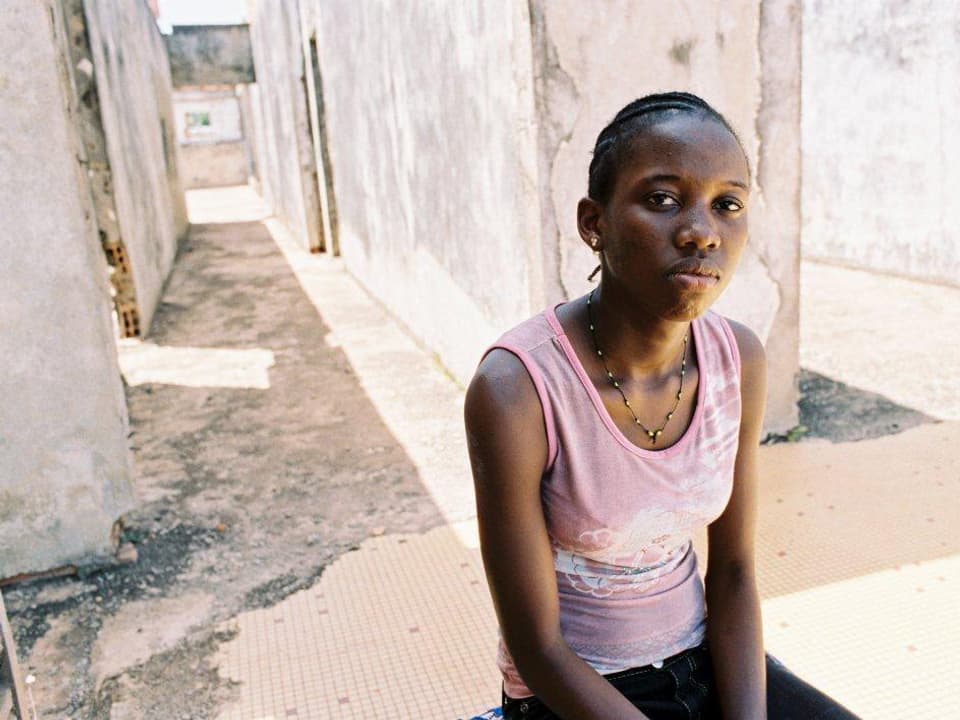 Ein junges schwarzes Mädchen steht in einer weiss getünchten Strasse und schaut nüchtern in die Kamera.