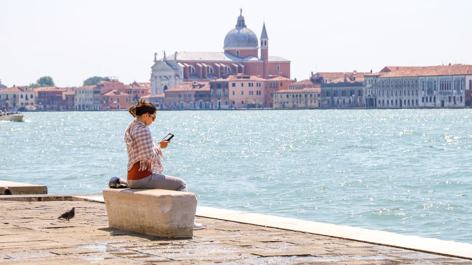 Eine junge Frau sitzt auf einer Steinbank am Meer. Im Hintergrund Venedig.