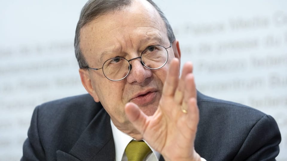 Mann mit Brille, seichtem grau-dunkelbraunem Haar und gelber Krawatte bei einer Pressekonferenz.