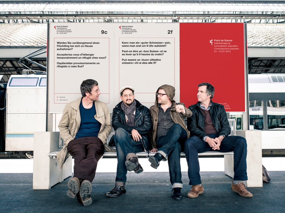 Vier Männer sitzen auf einer Bank im Bahnhof vor einem grossen Plakat.