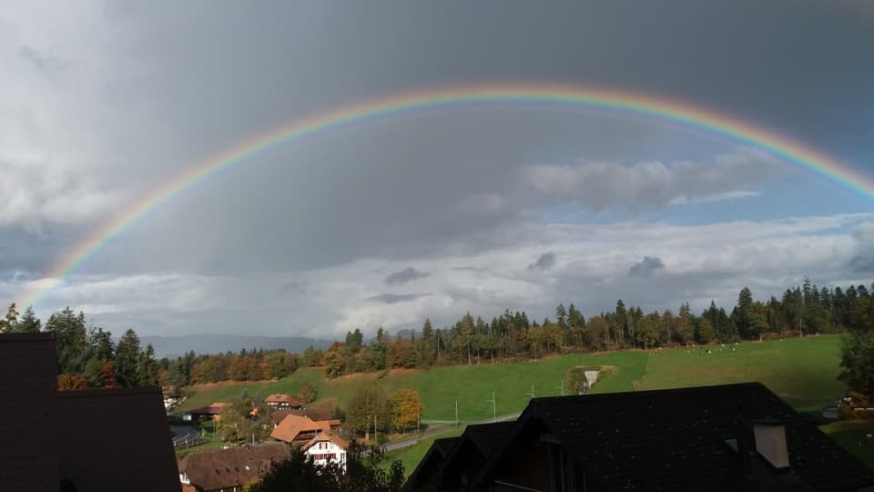 Regenbogen über Heimberg BE, darunter Wiese mit Bauernhaus und hinten Wald.