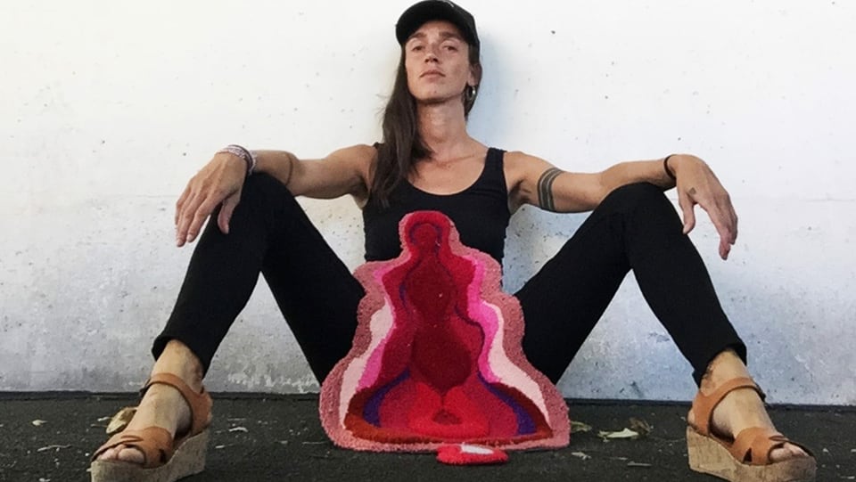 eine Frau sitzt an einer Wand, eine Plüsch-Vulva vor ihr