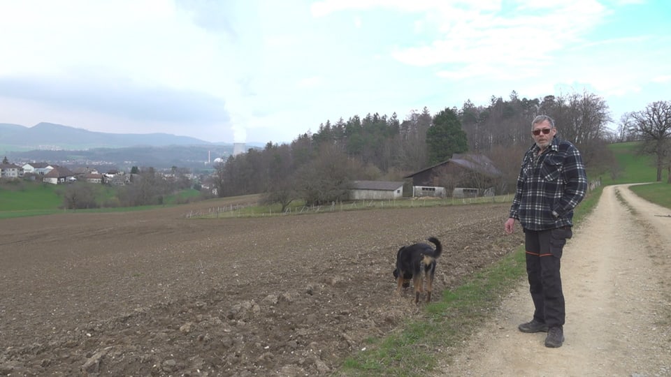 Ein Mann steht auf einem Feldweg mit seinem Hund, im Hintergrund ein Acker, Wald und Wiese. 