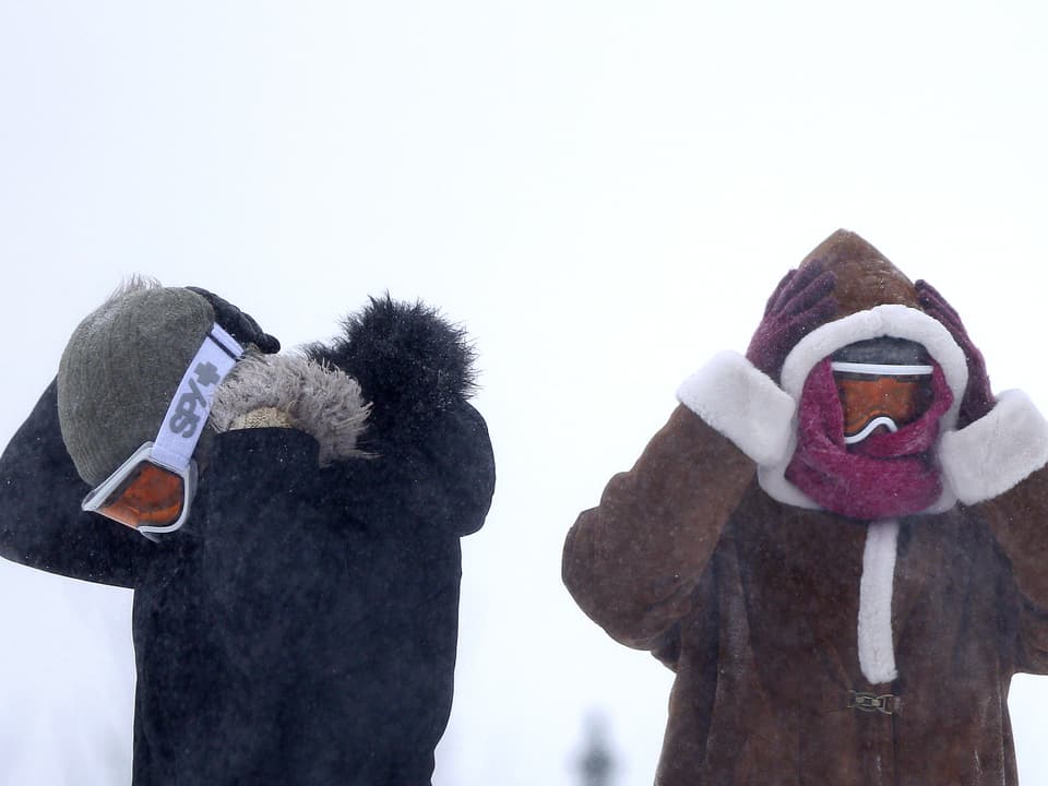 Zwei Spaziergänger im Schneesturm halten sich die Ohren unter ihren Winterkappen zu. Sie tragen zusätzlich Halstücher und Skibrillen.