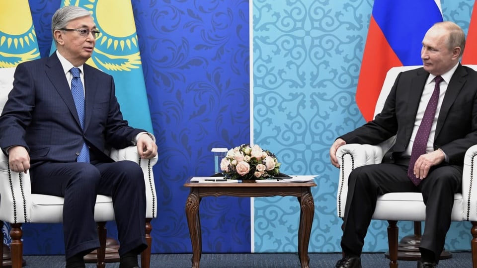 Tokajev und Putin sitzen auf zwei Sesseln nebeneinander, dahinter die Landesflaggen.