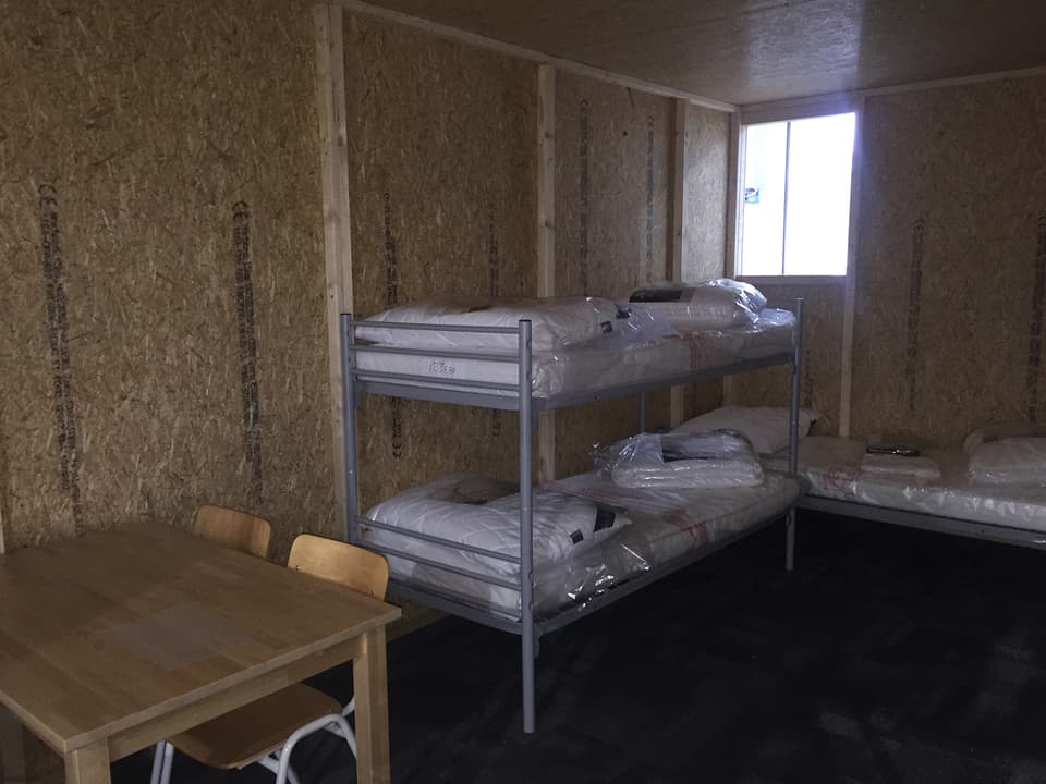 Stock-Bett und Bett in dunklem Raum mit Spanplattenwänden und einem kleinen Fenster im Hintergrund. 