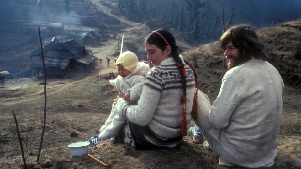 Ein Mann, eine Frau und ihr Baby sitzen auf einen Hügel mit Blick auf wenige, marode Gebäude.