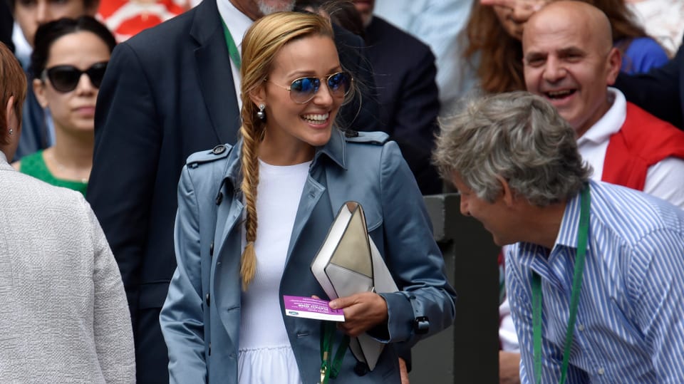 Jelena Djokovic trägt eine grosse Sonnenbrille, eine blaue Jacke und begrüsst jemanden im Publikum.