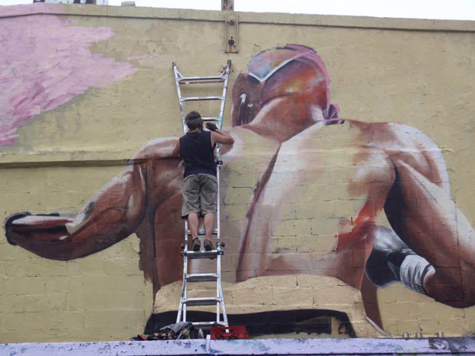 Ein Mann auf einer Leiter malt ein grosses Bild eines Boxers auf eineWand.
