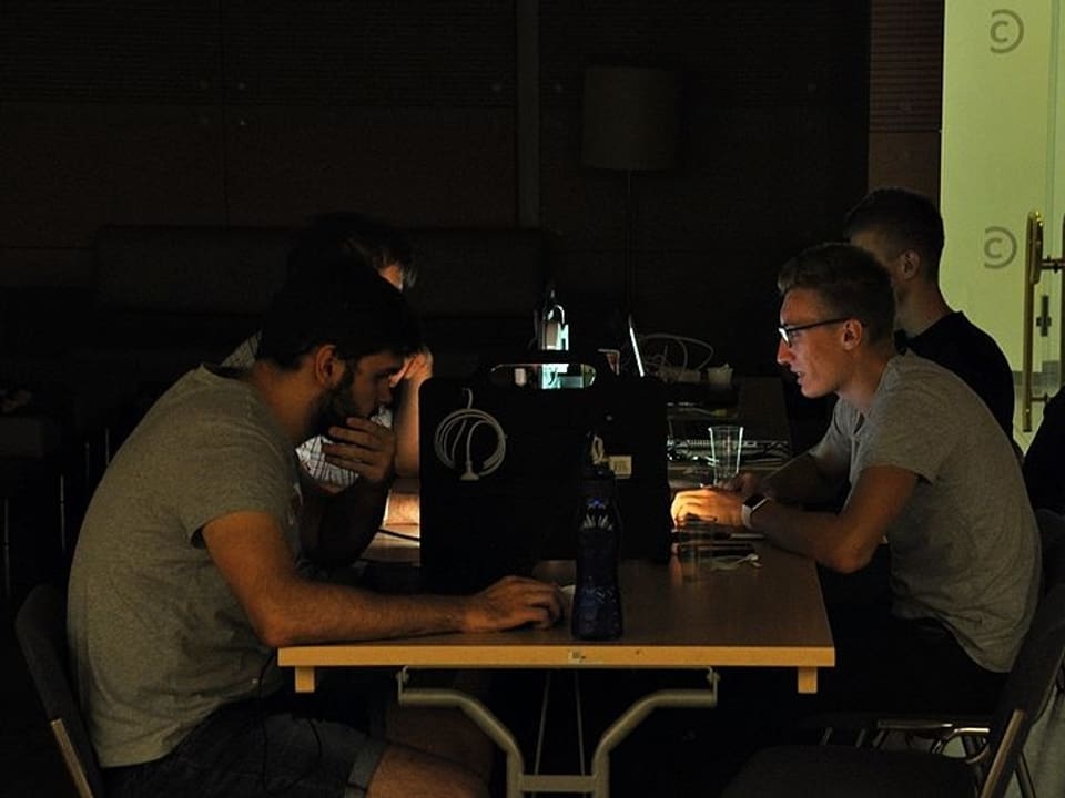 Gruppe junger Männder im Halbdunkel an einem Tisch mit verschiedenen Computern.