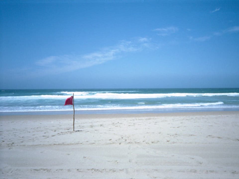 Strand, eine rote Fahne im Sand, dahinter blau das Meer.