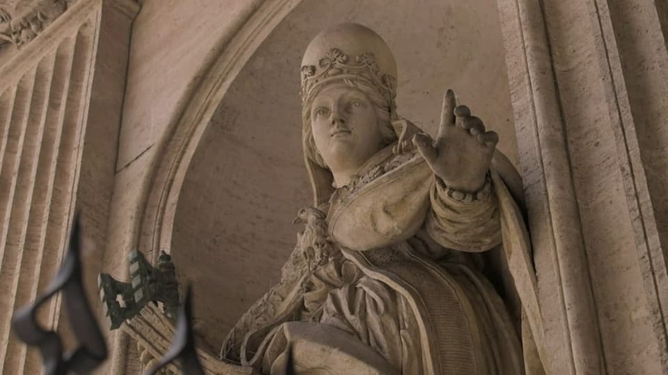 Foto einer Steinstatue. Eine Frau mit Tiara, hebt linke Hand, in einem steinernen Bogen.