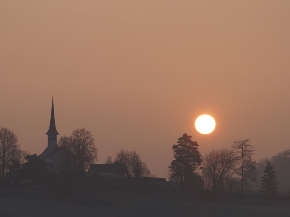 Bäume, Häuser und eine Kirche als Silhoutten. Über dem Horizont die Sonne durch den leicht rötlichen Nebel.