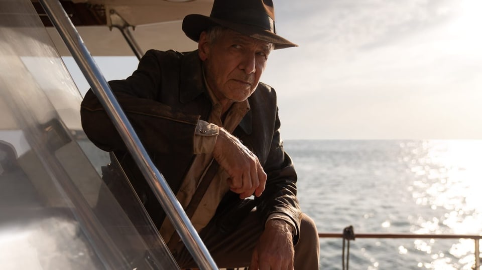 Grau melierter Mann mit Hut sitzt auf einem Boot in der Hocke und schaut skeptisch.
