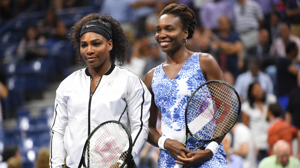 Serena und Venus Williams lächeln am Netz in die Kameras.