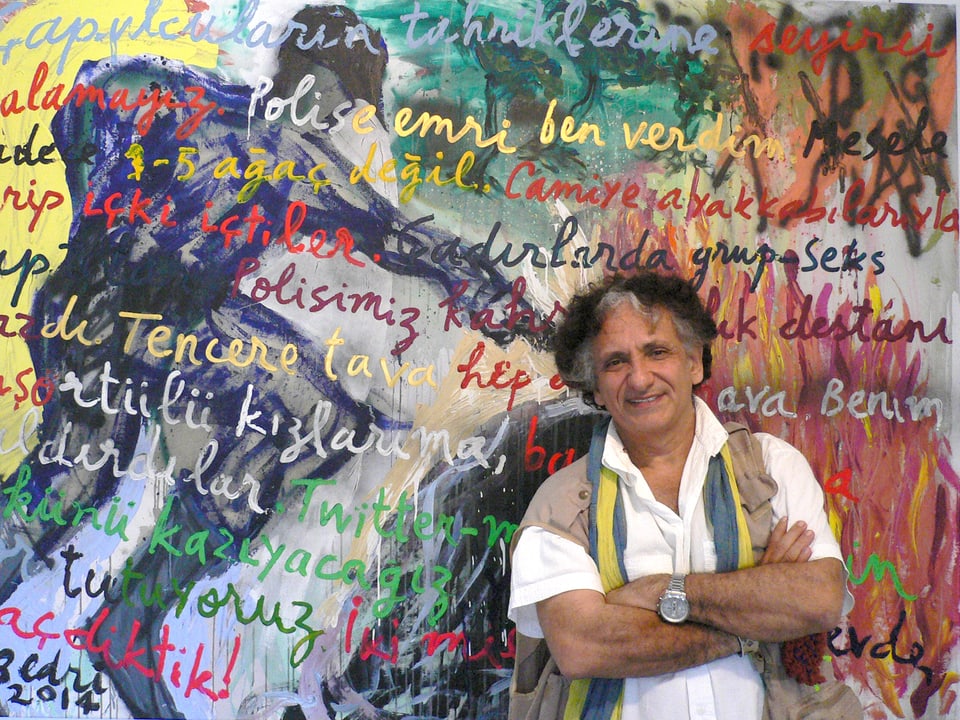 Ein Künstler steht vor seinem Gemälde – drauf sind verschiedene handgeschriebene, farbige Sprüche.