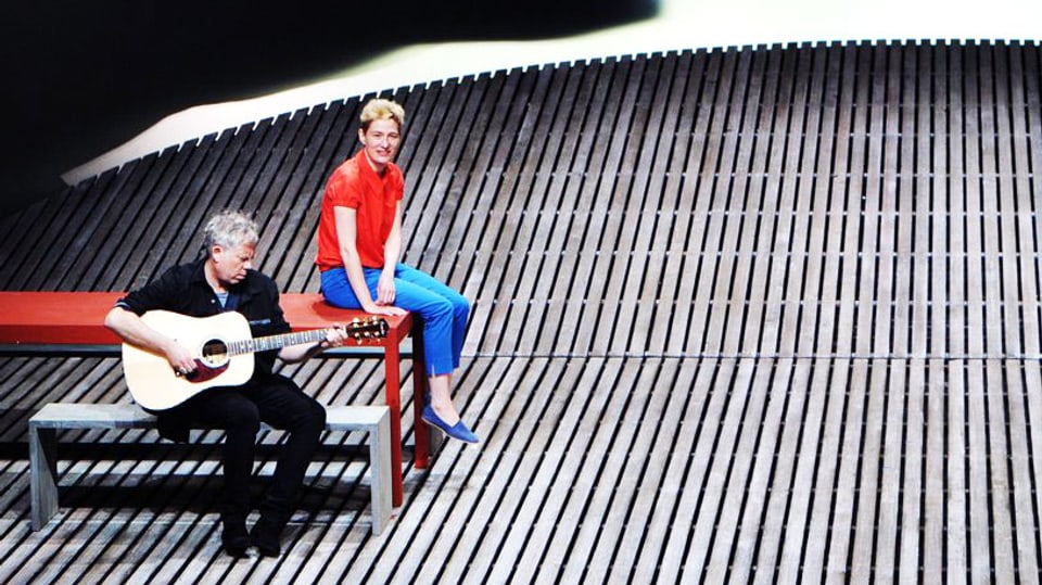 Eine Mann spielt Gitarre auf einer Bühne. Neben ihm sitzt eine Frau.