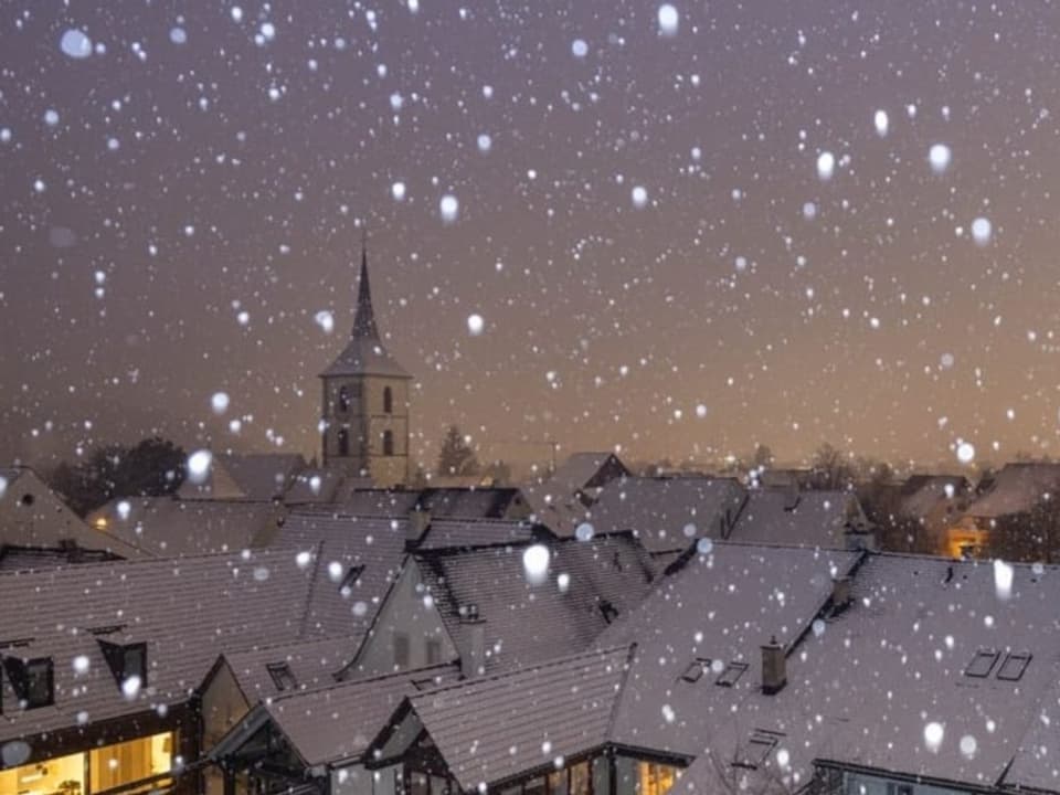 Dorf mit verschneiten Häusern, auch durch die Luft tanzen weitere Schneeflocken im ersten Morgenlicht.
