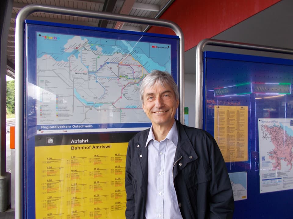 Brühlmann hat mittellange graue Haare und steht am Bahnhof Amriswil vor einem gelben Fahrplan.