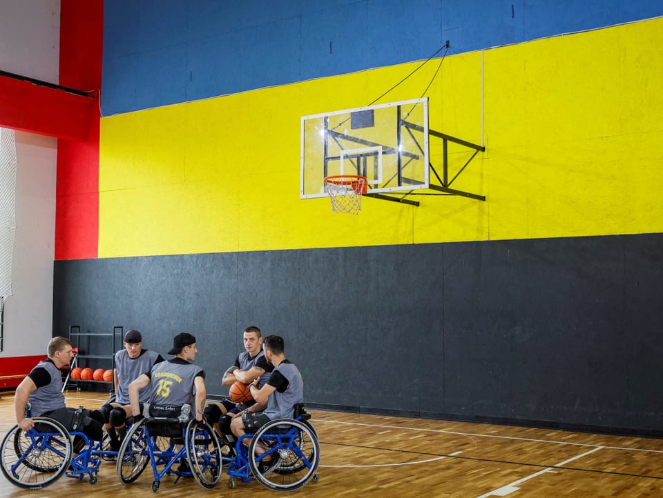 Fünf Männer im Rollstuhl in einer Basketball-Halle. Die Wand dahinter trägt die Farben der ukrainischen Flagge.
