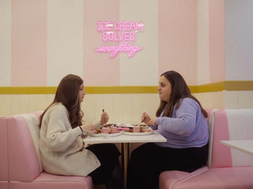 Jochewed und ihre Freundin Gitty sitzen in einer rosafarbenen Eisdiele und essen Waffeln.