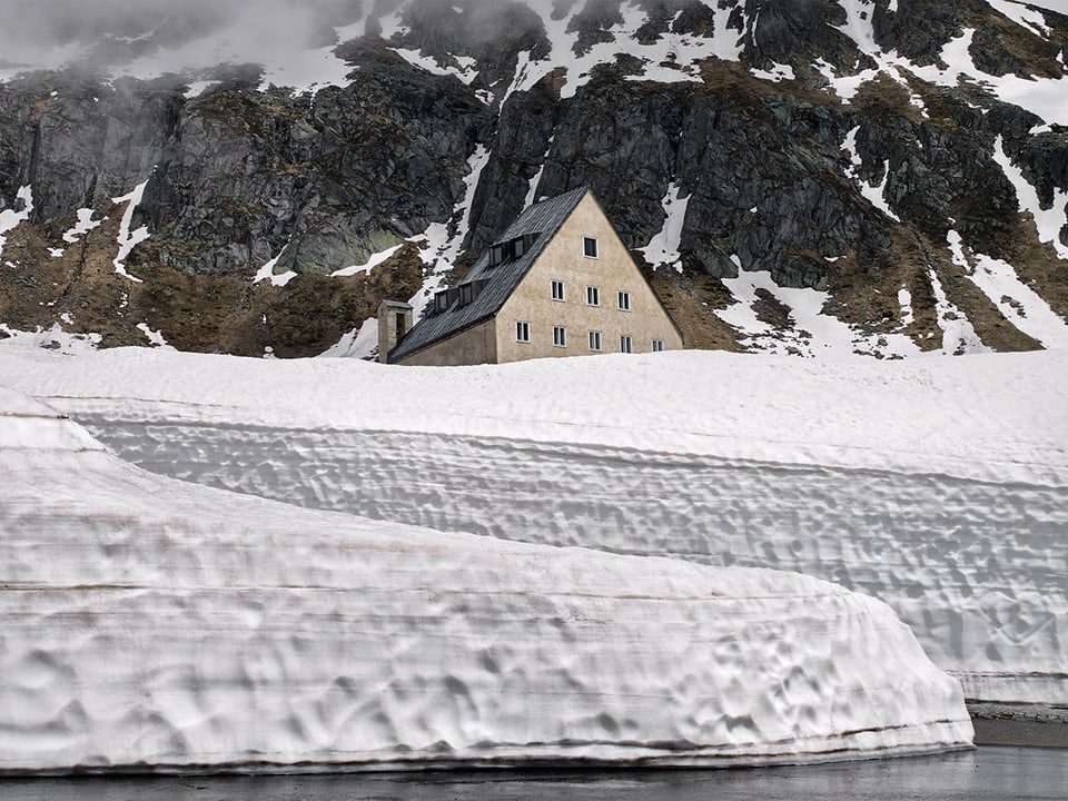 Haus vor Bergwand, im Vordergrund ein Schneewall.