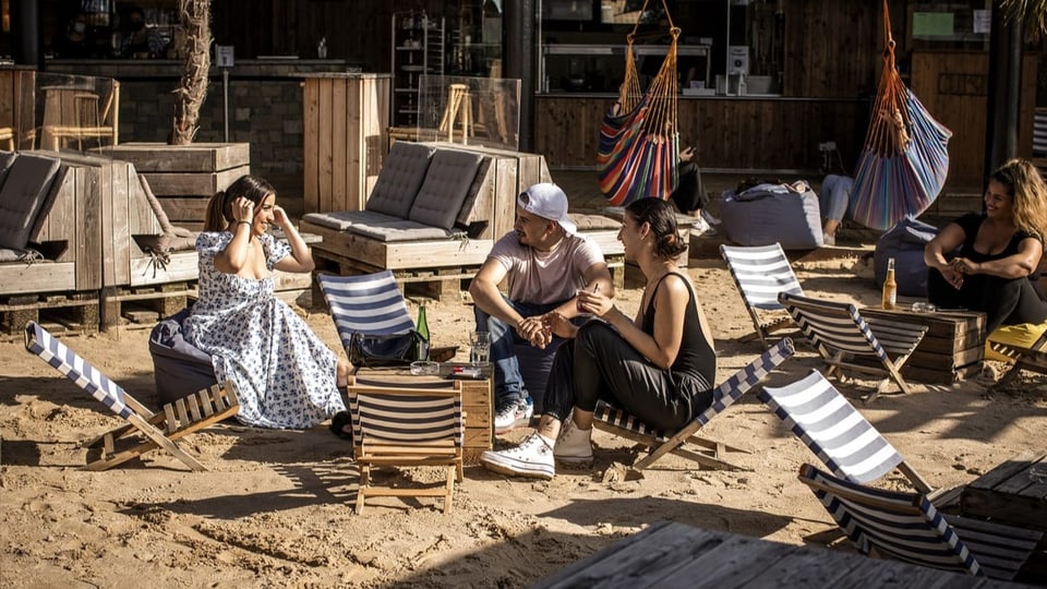 Menschen sitzen im Sand auf Liegestühlen, im Hintergrund eine Bar aus Holz