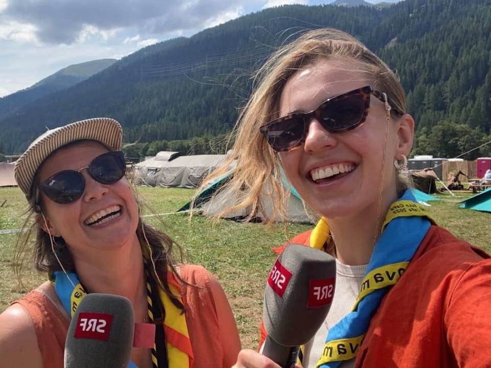 Zwei Frauen halten ein Mikrofon und lachen in die Kamera. Sie sind auf einem Zeltplatz.