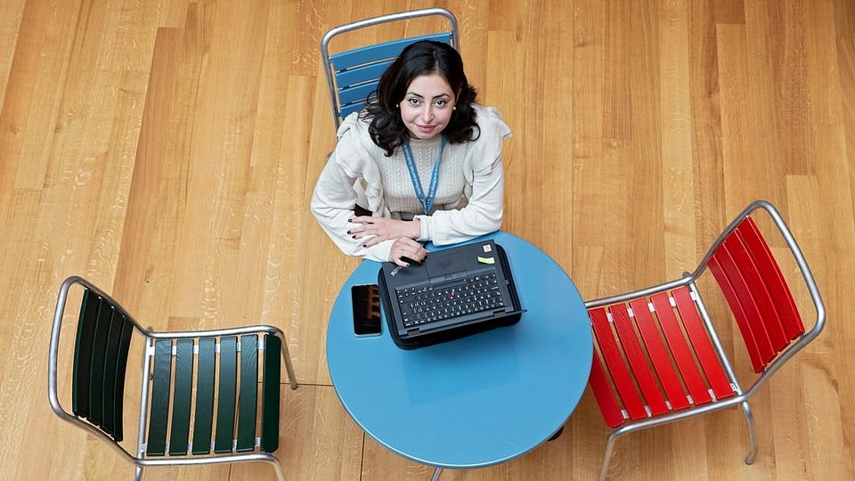 Eine Frau sitzt an einem kleinen blauen Tischchen mit Laptop drauf und blickt nach oben.