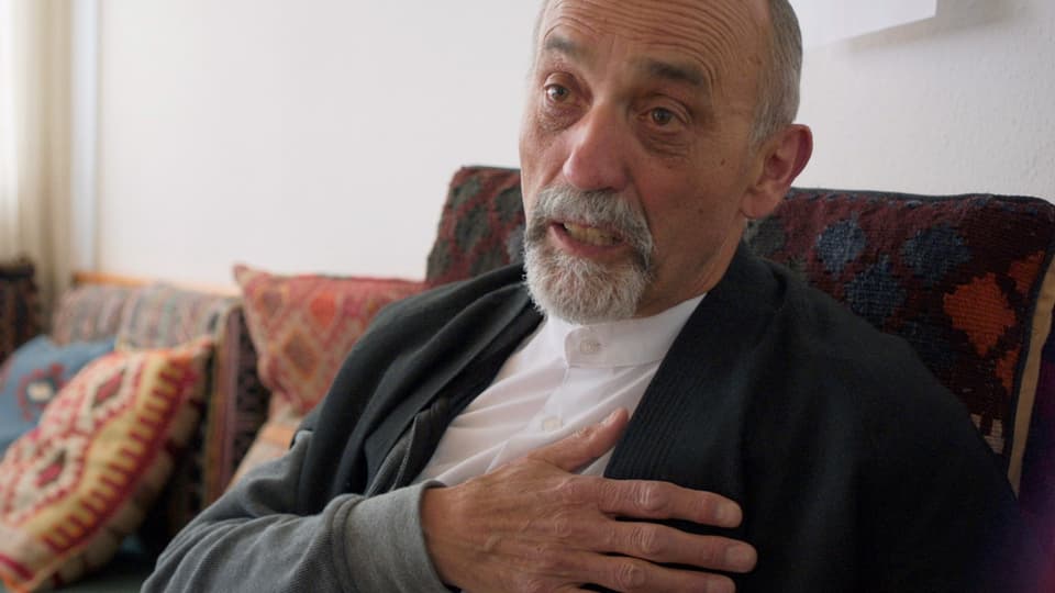 Ein älterer Mann mit grauem Bart sitzt auf einem Sofa mit bunten Kissen.