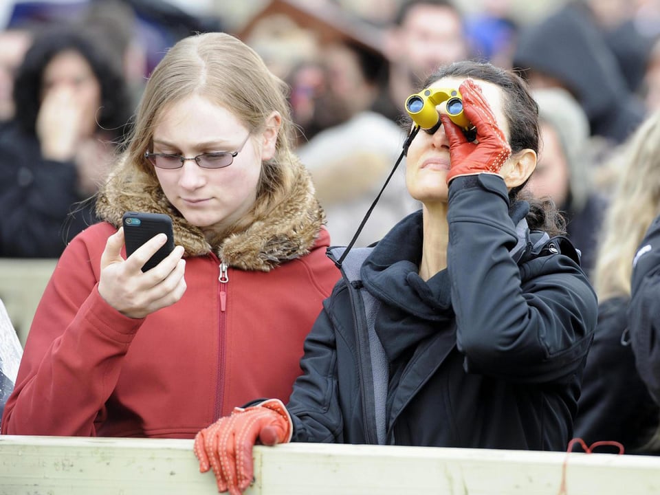 Frau schaut mit Opernglas in Richtung Schornstein, während die Frau neben ihr ihr Smartphone betrachtet.